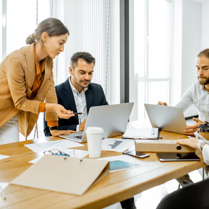 Team of professionals working together at a conference table with laptops, documents, and coffee cups during a business meeting in a bright office.