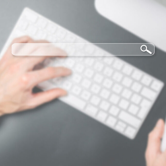 Blurred image of a person typing on a white keyboard with a transparent search bar and magnifying glass icon overlayed on the screen.
