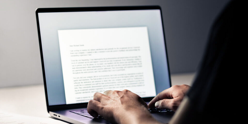 Close-up of hands typing on a laptop keyboard with a document open on the screen in a dimly lit workspace.