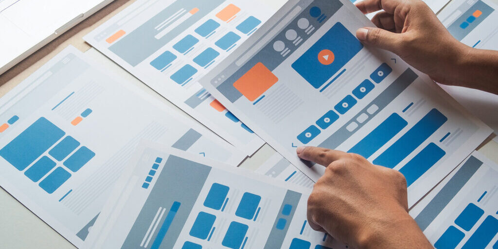 Hands holding a printed website wireframe layout among multiple user interface design mockups spread out on a table during the planning phase of a web project.