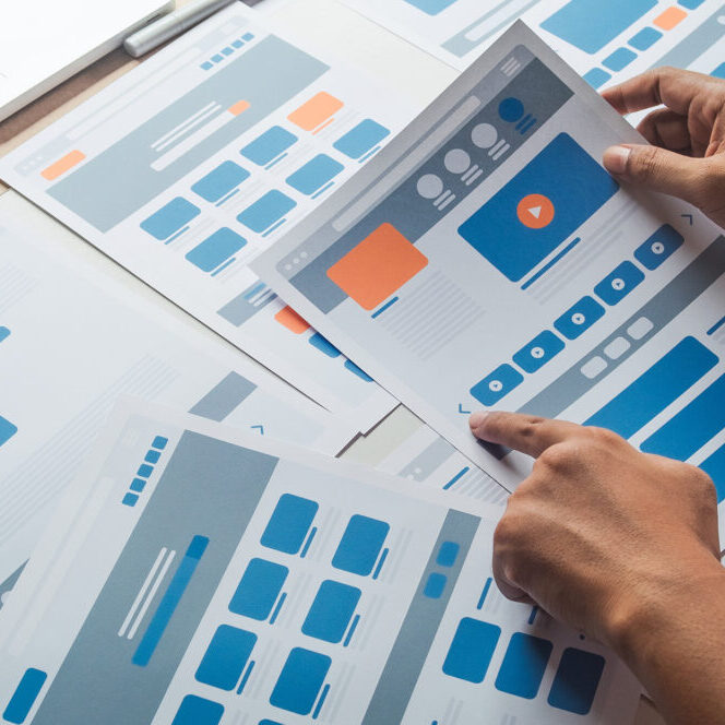 Hands holding a printed website wireframe layout among multiple user interface design mockups spread out on a table during the planning phase of a web project.