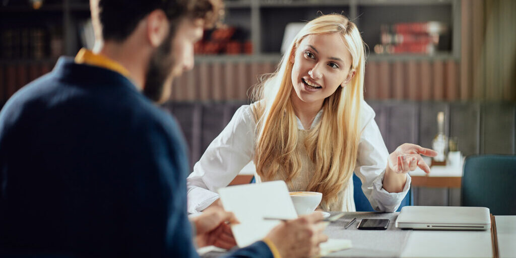 Woman smiling and gesturing while having a conversation with a man in a casual meeting setting, with notebooks, a tablet, and a coffee cup on the table.