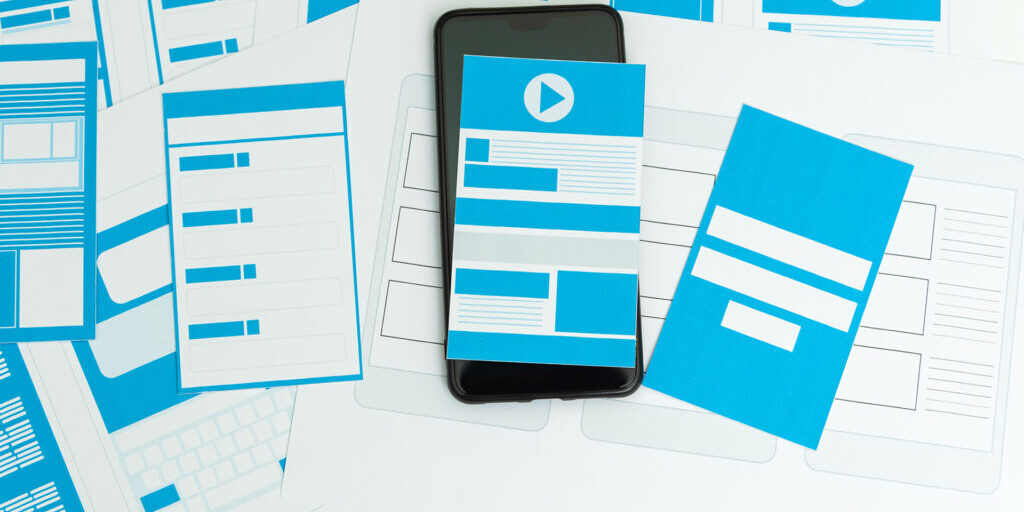 Smartphone placed on top of wireframe design sketches and blue layout mockups for mobile and web interfaces, illustrating app or website prototyping.