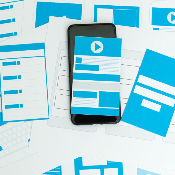 Smartphone placed on top of wireframe design sketches and blue layout mockups for mobile and web interfaces, illustrating app or website prototyping.