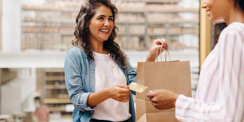 Smiling woman handing a credit card to a cashier while holding a shopping bag at a retail store counter.
