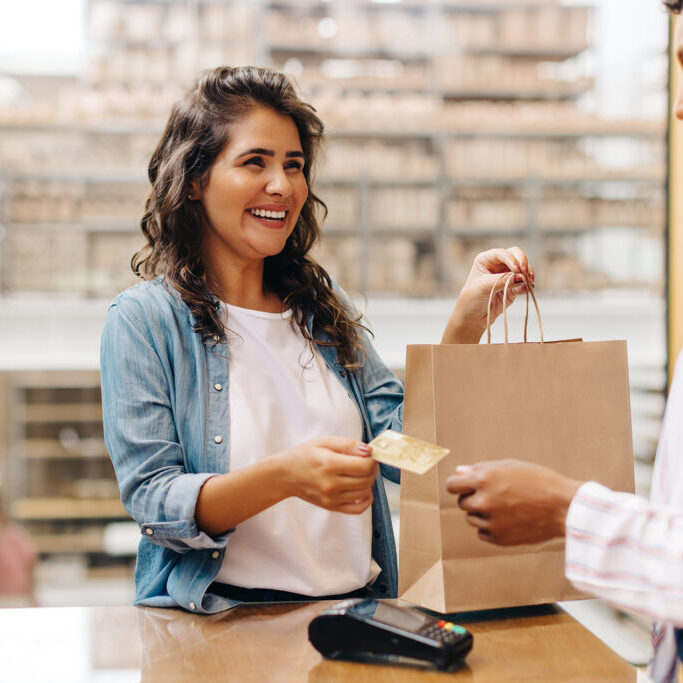Smiling woman handing a credit card to a cashier while holding a shopping bag at a retail store counter.