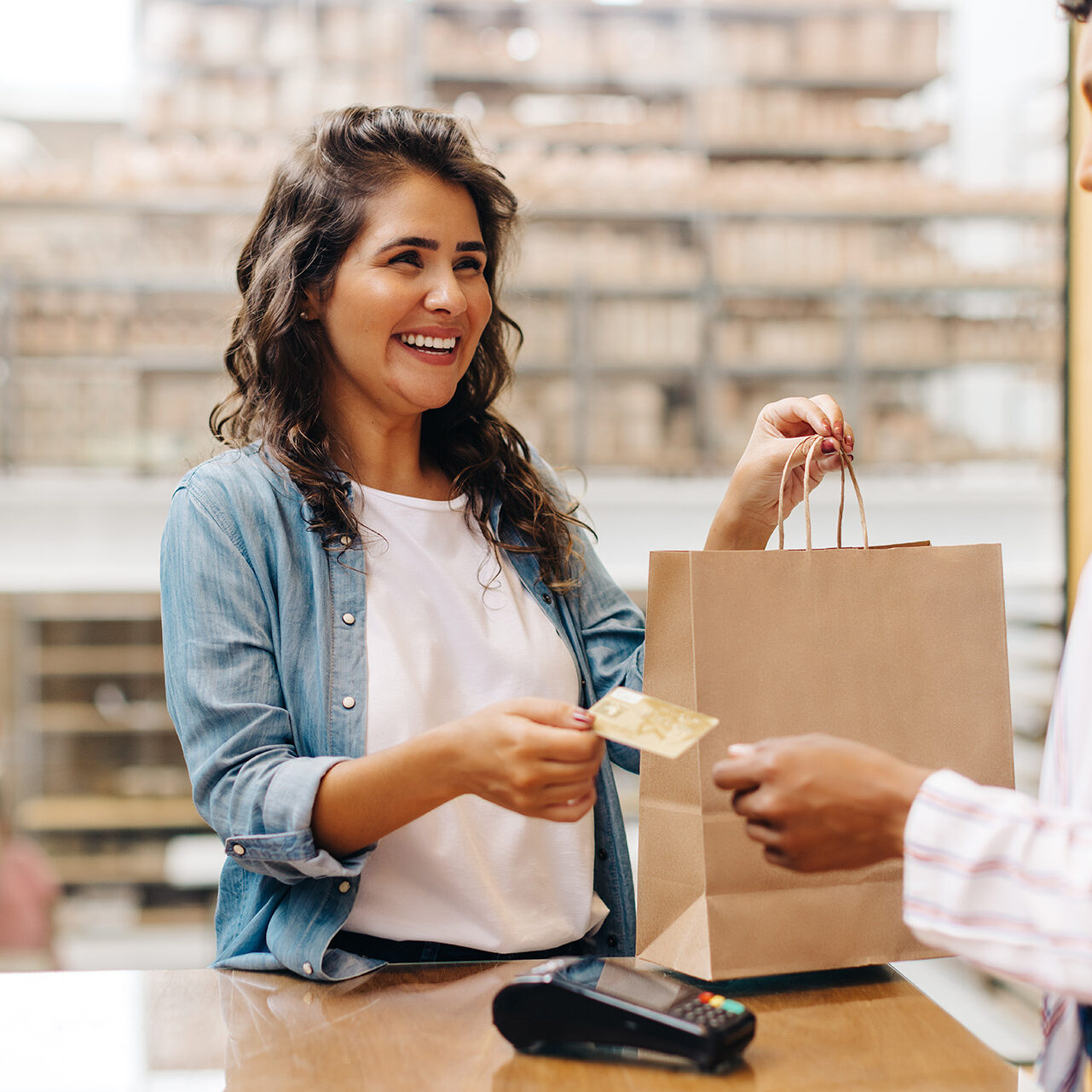 Smiling woman handing a credit card to a cashier while holding a shopping bag at a retail store counter.