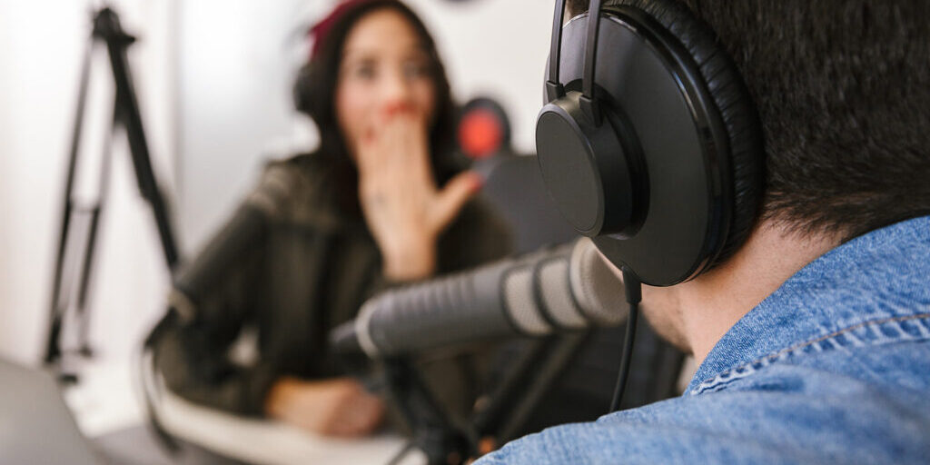 Close-up of a man wearing headphones speaking into a microphone during a podcast recording, with a woman in the background reacting with a surprised expression.
