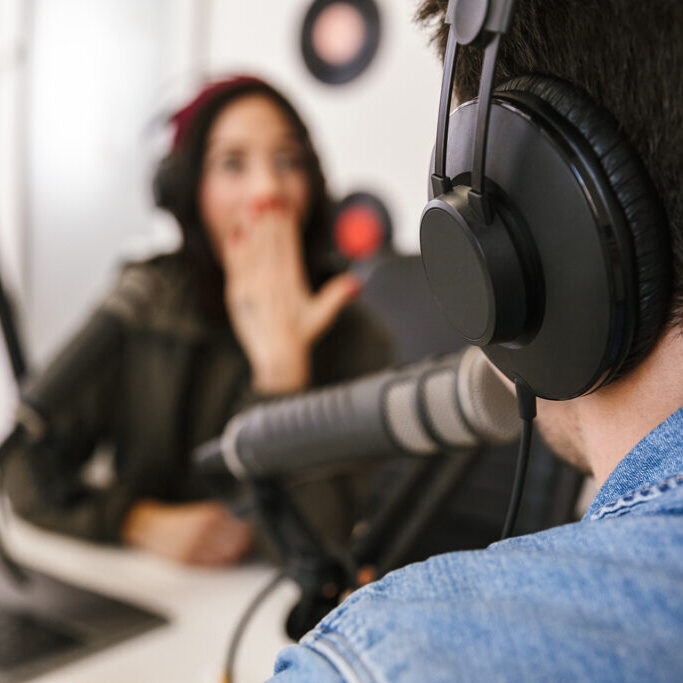 Close-up of a man wearing headphones speaking into a microphone during a podcast recording, with a woman in the background reacting with a surprised expression.