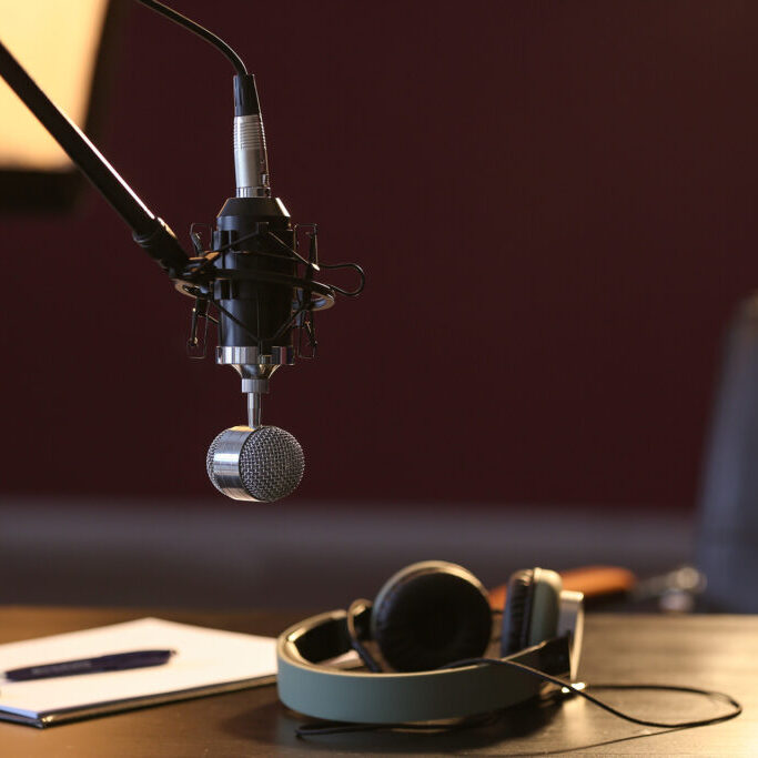 Professional studio microphone suspended on a boom arm above a desk with a pair of headphones and a notebook, set up for podcasting or voice recording.