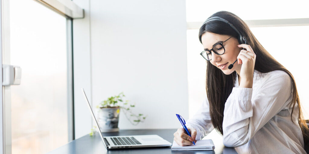 Woman wearing a headset and glasses taking notes in a notebook while attending a video call on her laptop in a bright office space.
