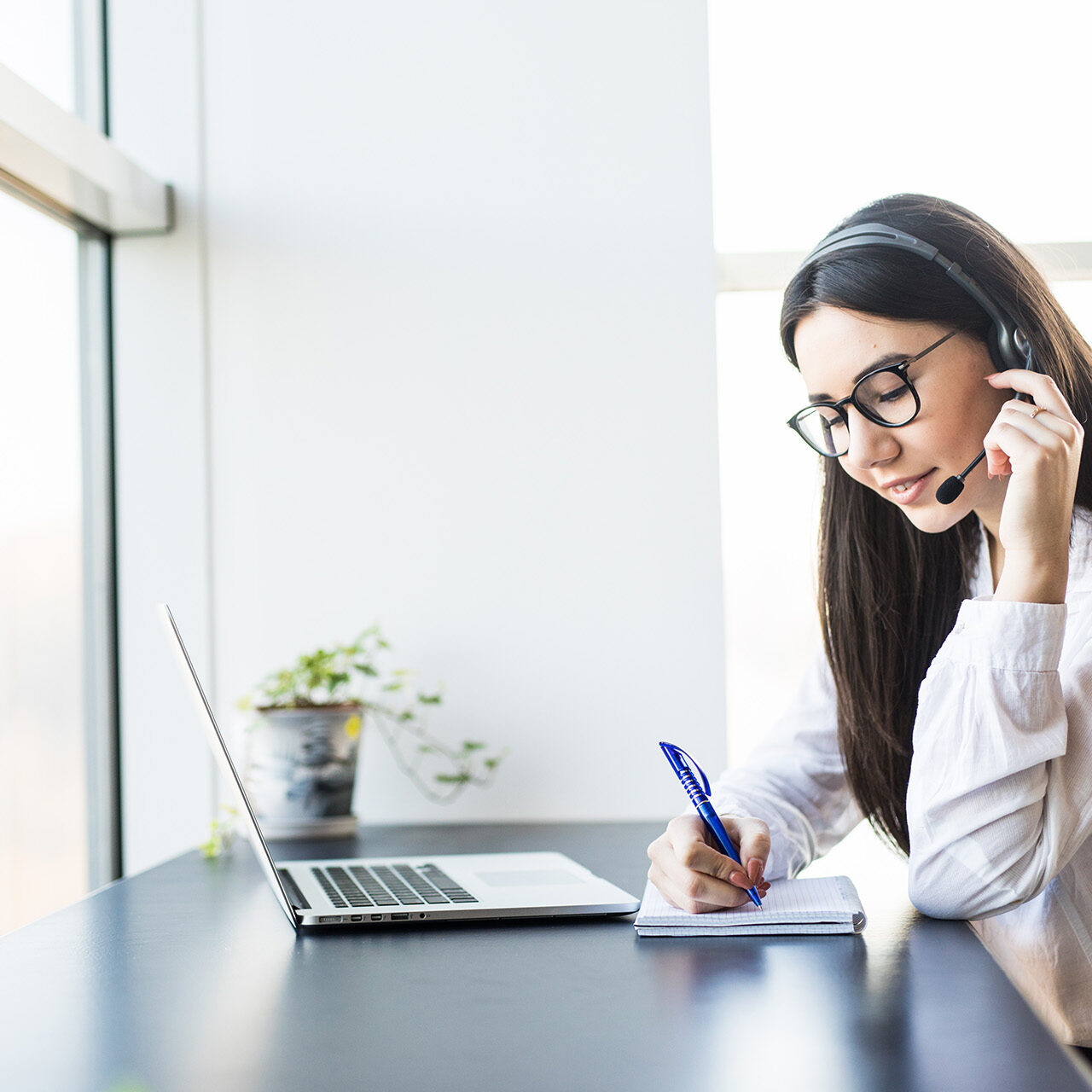 Woman wearing a headset and glasses taking notes in a notebook while attending a video call on her laptop in a bright office space.