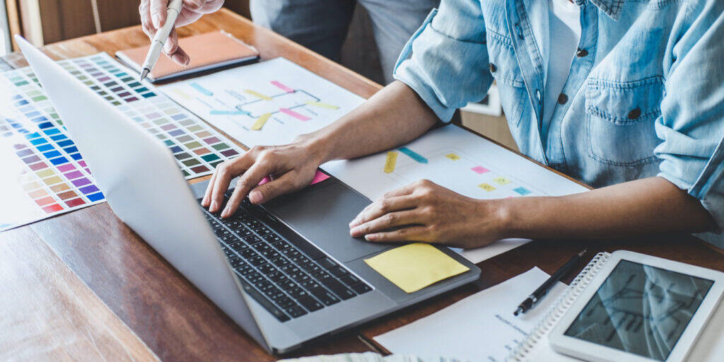 Close-up of a designer's hands working on a laptop surrounded by color swatches, wireframes, and printed layout designs during a creative planning session.