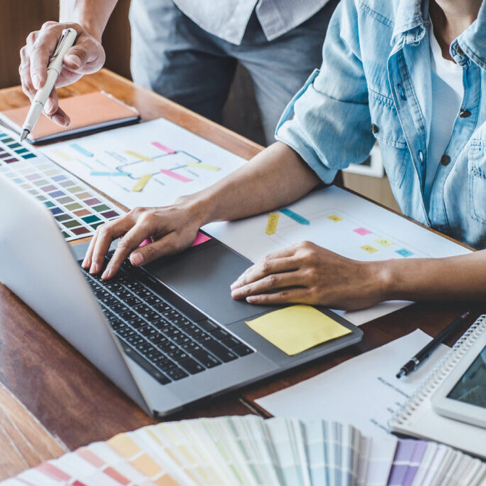 Close-up of a designer's hands working on a laptop surrounded by color swatches, wireframes, and printed layout designs during a creative planning session.