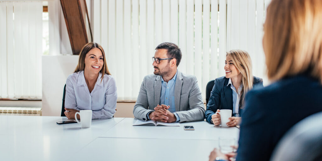 Group of business professionals sitting around a conference table, smiling and engaging in a meeting with notebooks, coffee mugs, and a smartphone on the table.
