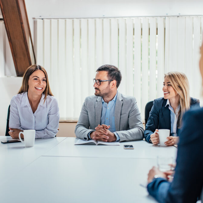 Group of business professionals sitting around a conference table, smiling and engaging in a meeting with notebooks, coffee mugs, and a smartphone on the table.