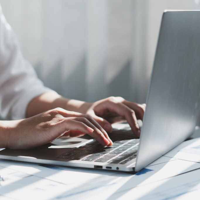 Close-up of a person typing on a laptop keyboard with sunlight streaming through window blinds in the background.