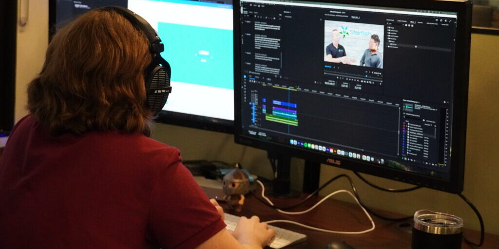 Person wearing headphones editing a video on a dual-monitor setup, with a video timeline and preview displayed on one screen and a webpage on the other.