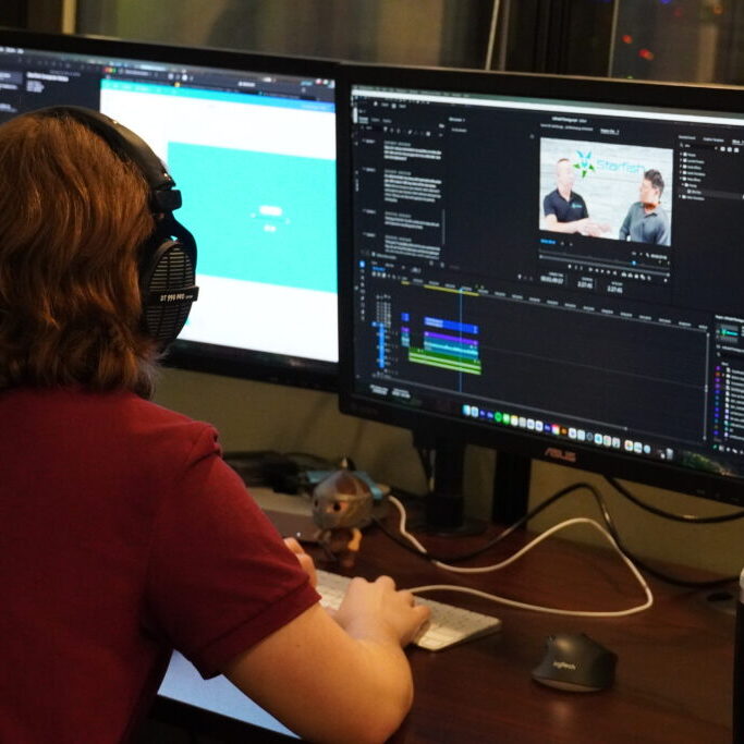 Person wearing headphones editing a video on a dual-monitor setup, with a video timeline and preview displayed on one screen and a webpage on the other.