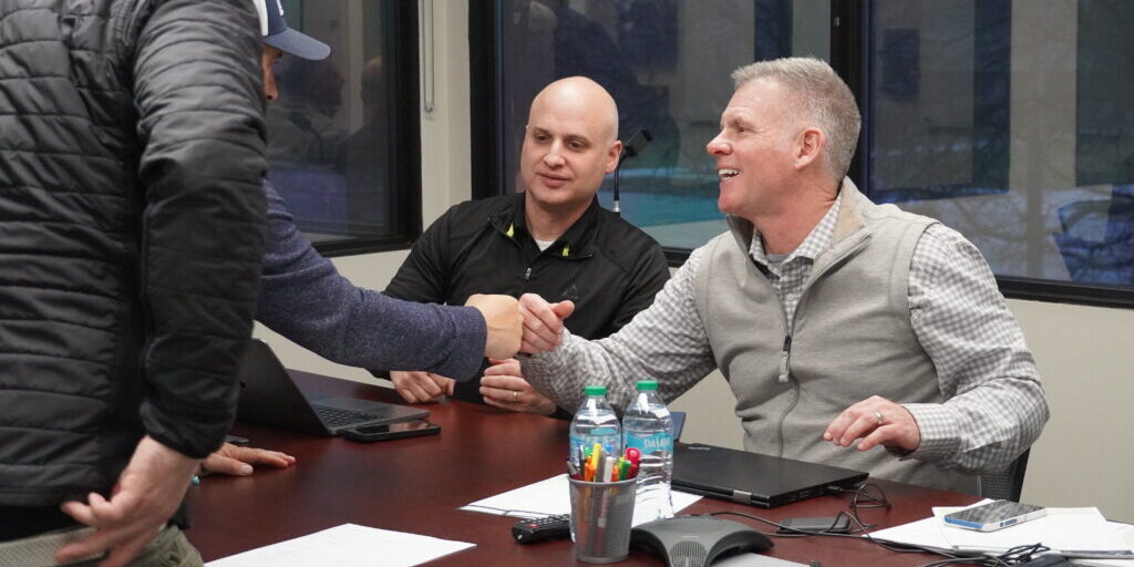 Two men shaking hands across a conference table during a business meeting, with another man observing and laptops, papers, and water bottles on the table.