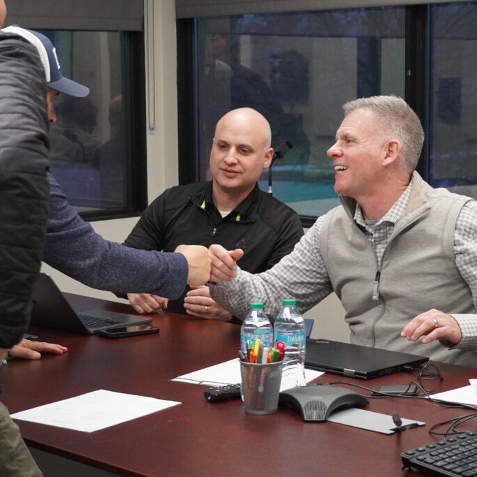 Two men shaking hands across a conference table during a business meeting, with another man observing and laptops, papers, and water bottles on the table.