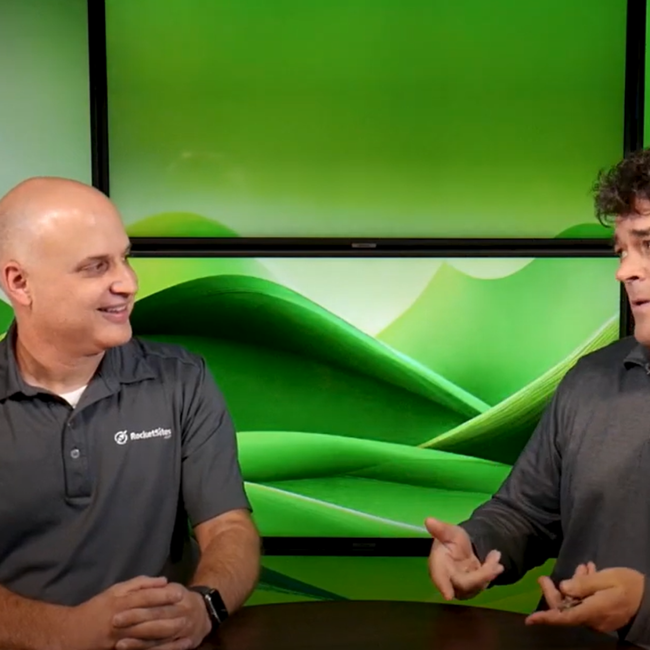 Two men sitting at a table having a conversation in front of a green abstract background displayed on large screens.