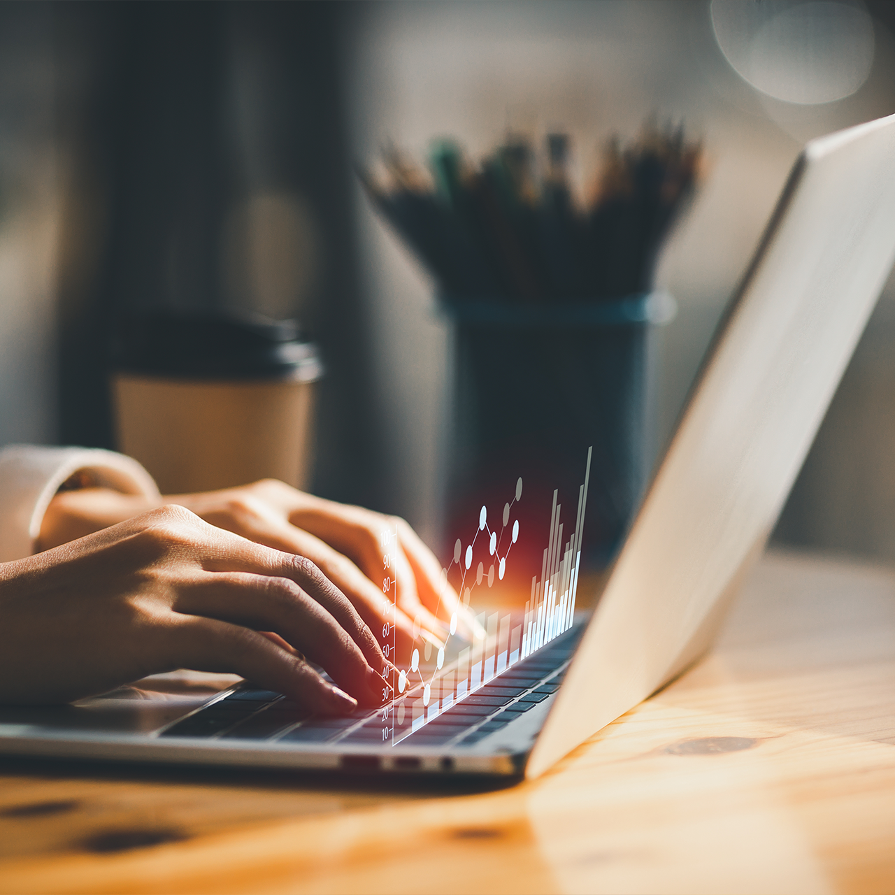 Close-up of hands typing on a laptop with digital data graphs overlaid, a coffee cup and pencil holder blurred in the background on a wooden desk